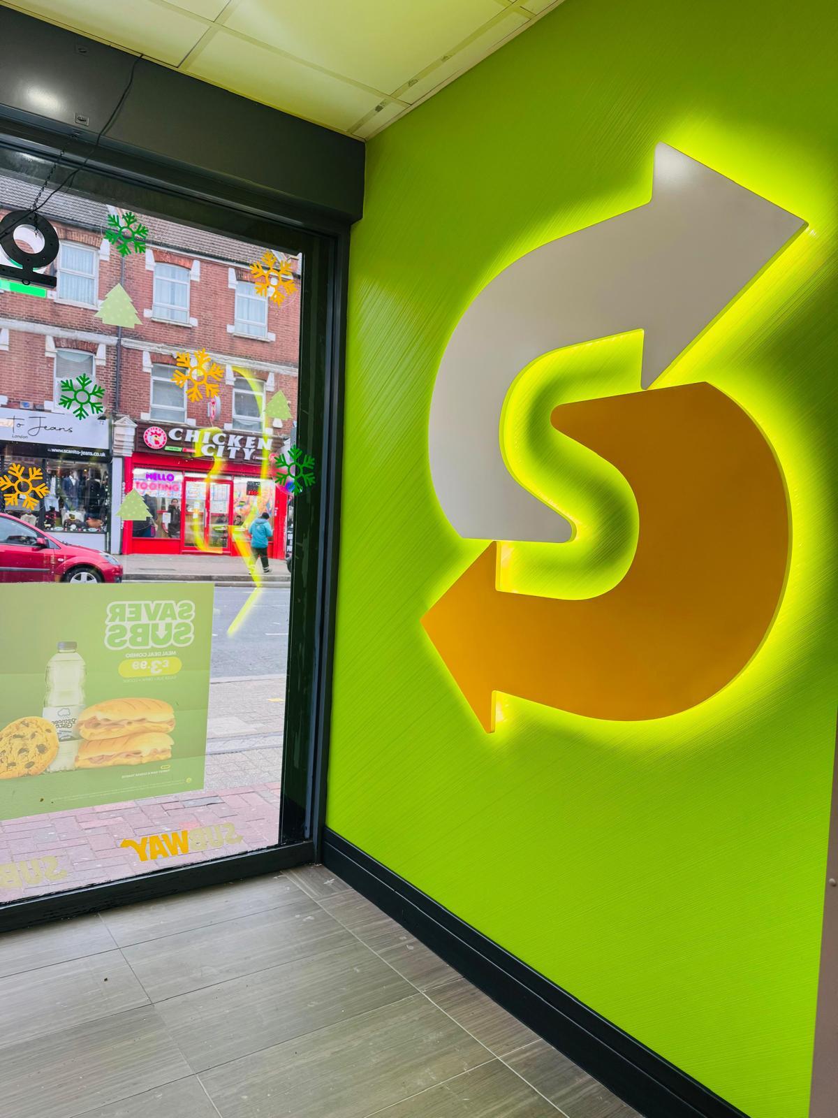 Illuminated Subway arrow logo on a green wall, with a shopfront window looking out onto the street.