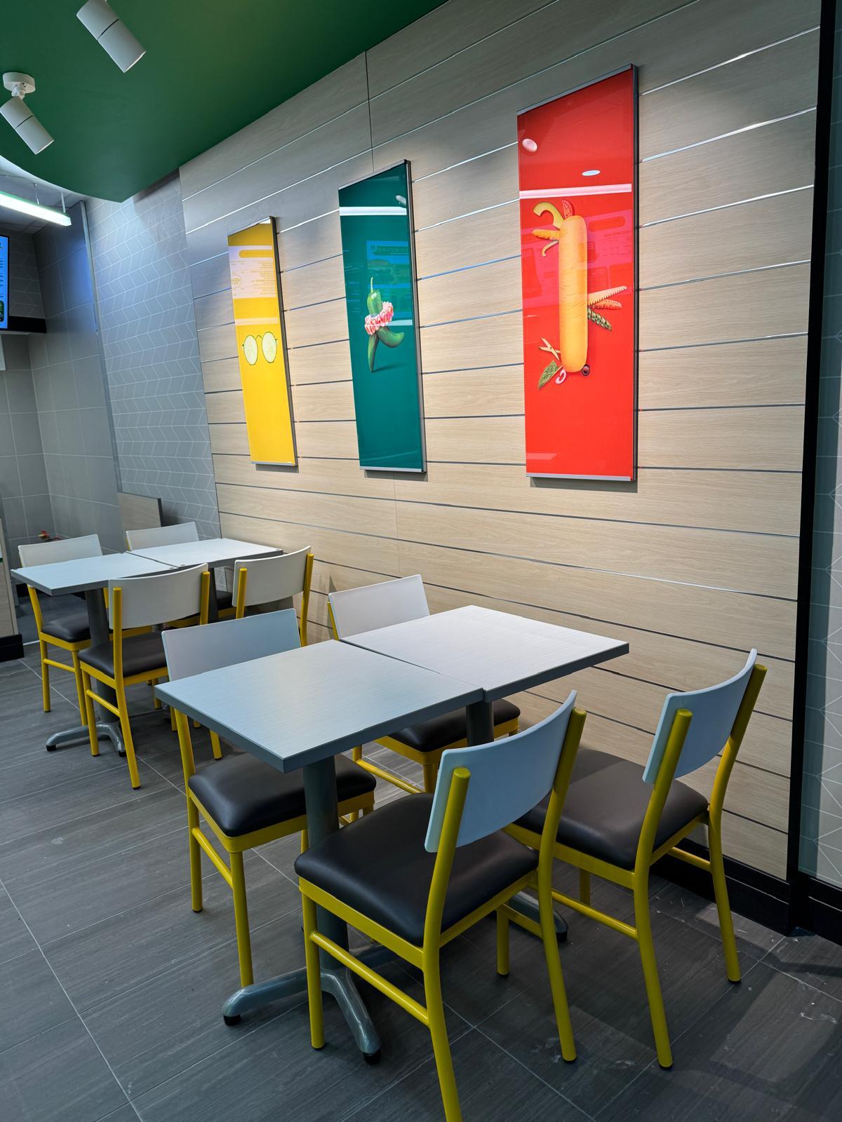 Subway dining area with colourful vegetable-themed wall art and seating with yellow-framed chairs.