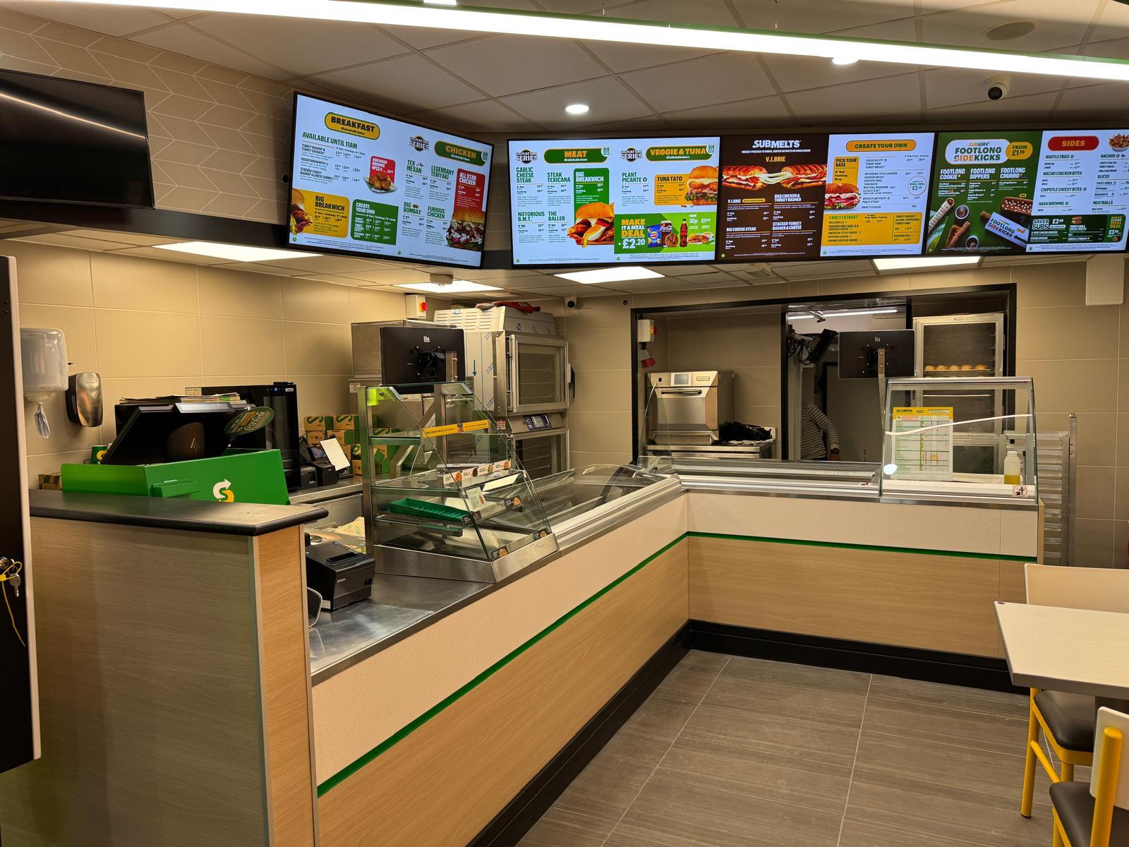 Sandwich preparation counter inside a Subway restaurant, with digital menu boards showing the breakfast and meat ranges.