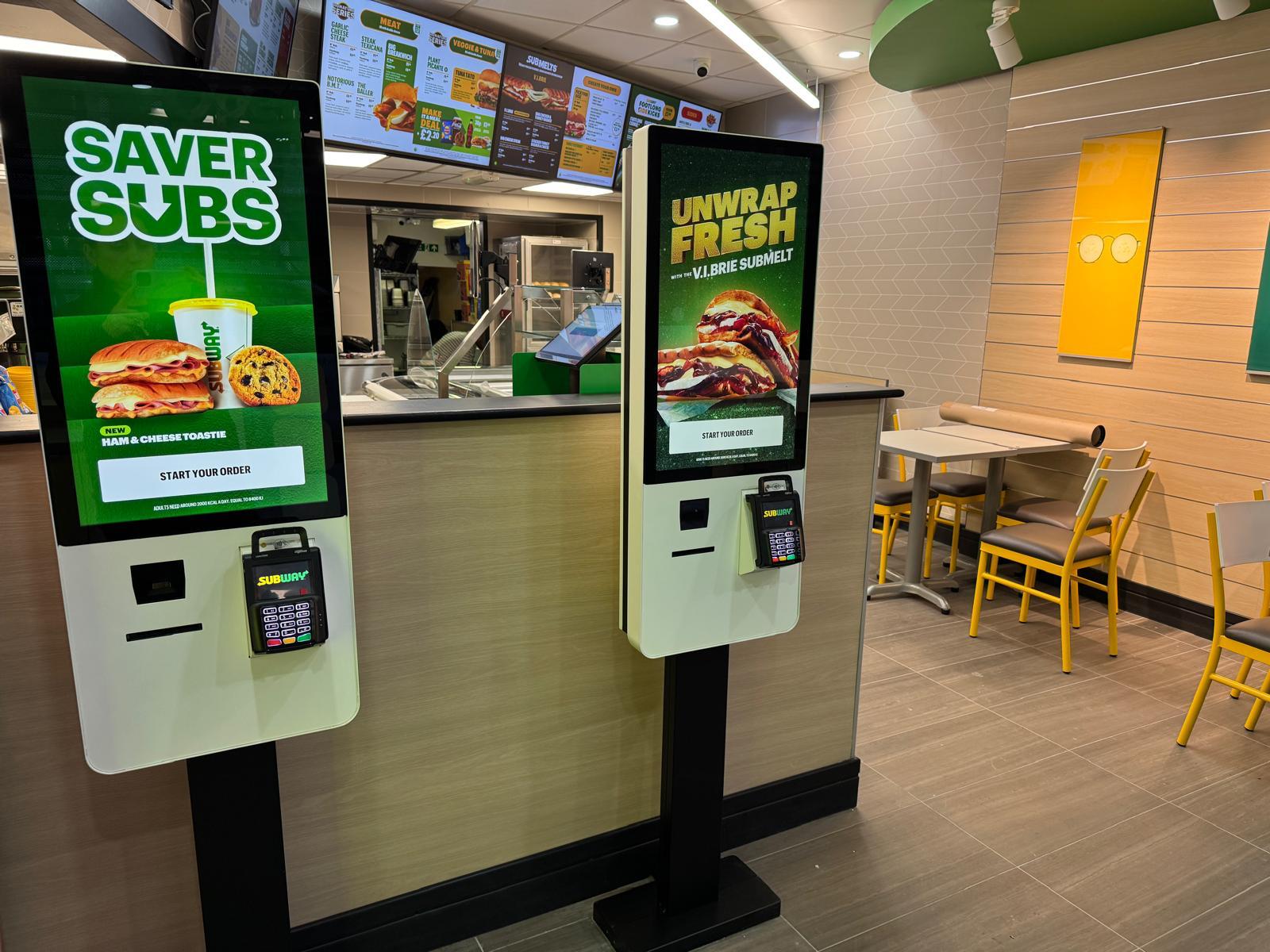 Self-service ordering kiosks inside a Subway restaurant, with digital menu boards and dining seating in the background.