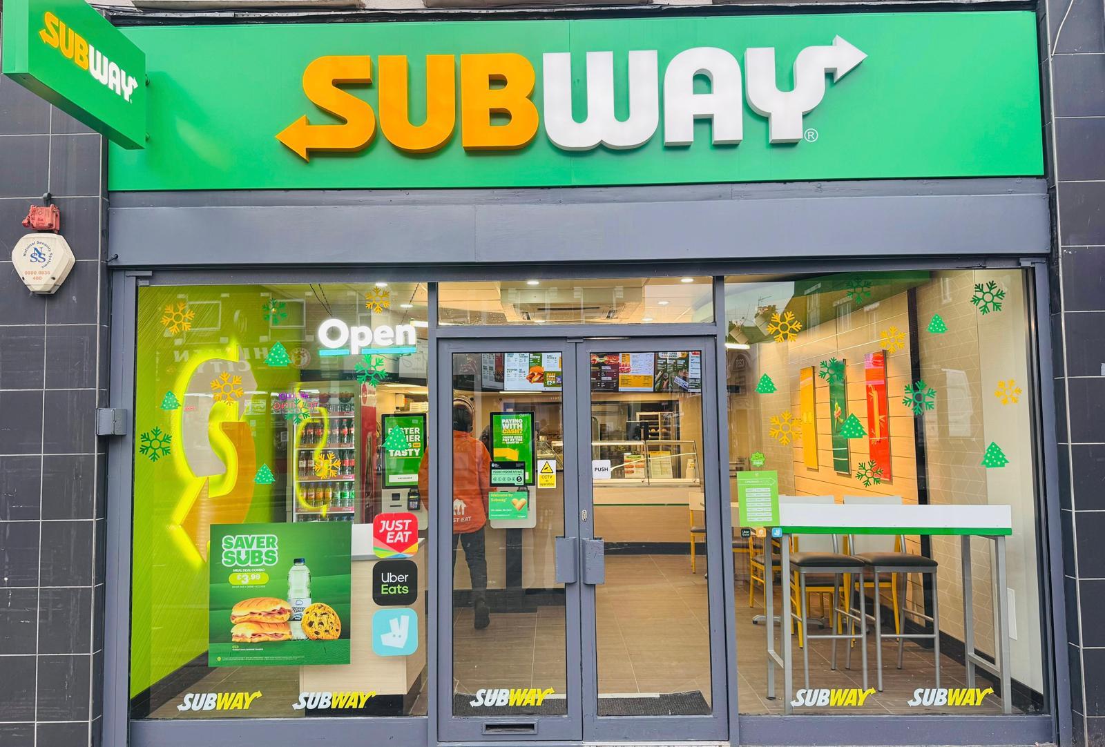 Exterior of a Subway shopfront with illuminated signage, an 'Open' sign, and delivery service stickers for Just Eat and Uber Eats.