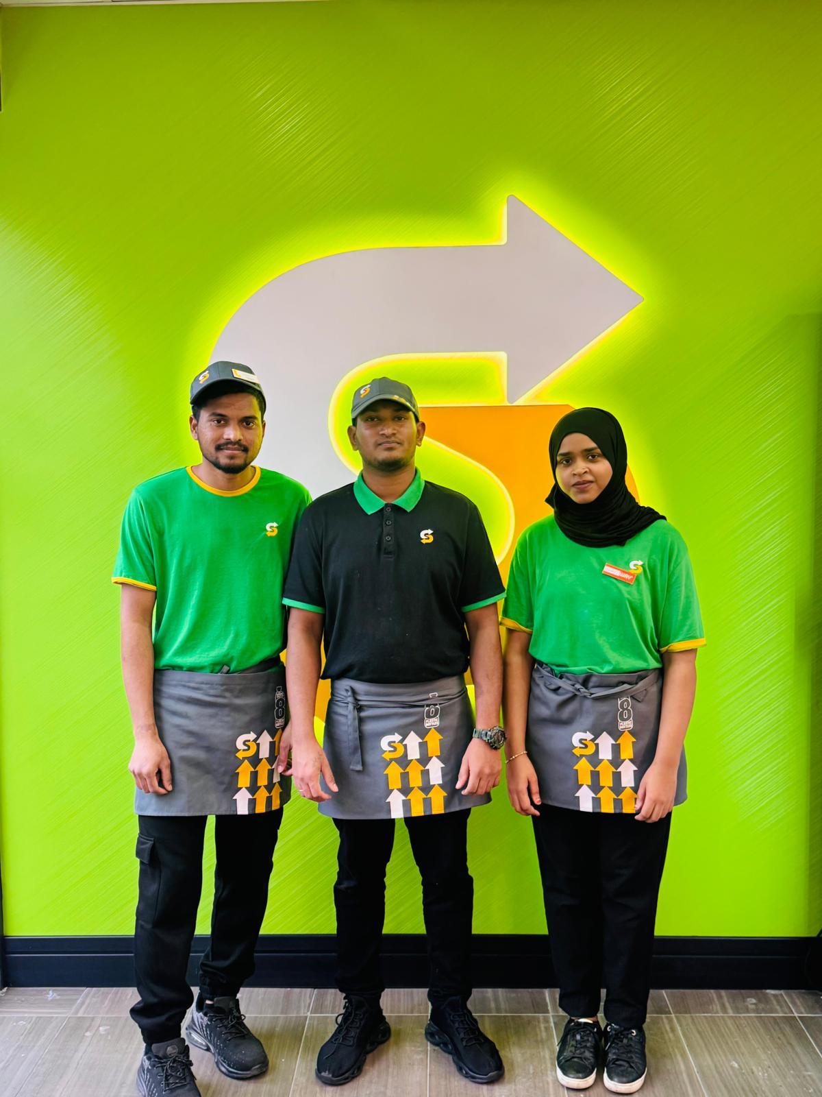 Three Subway staff members in branded uniforms standing in front of an illuminated Subway arrow wall.