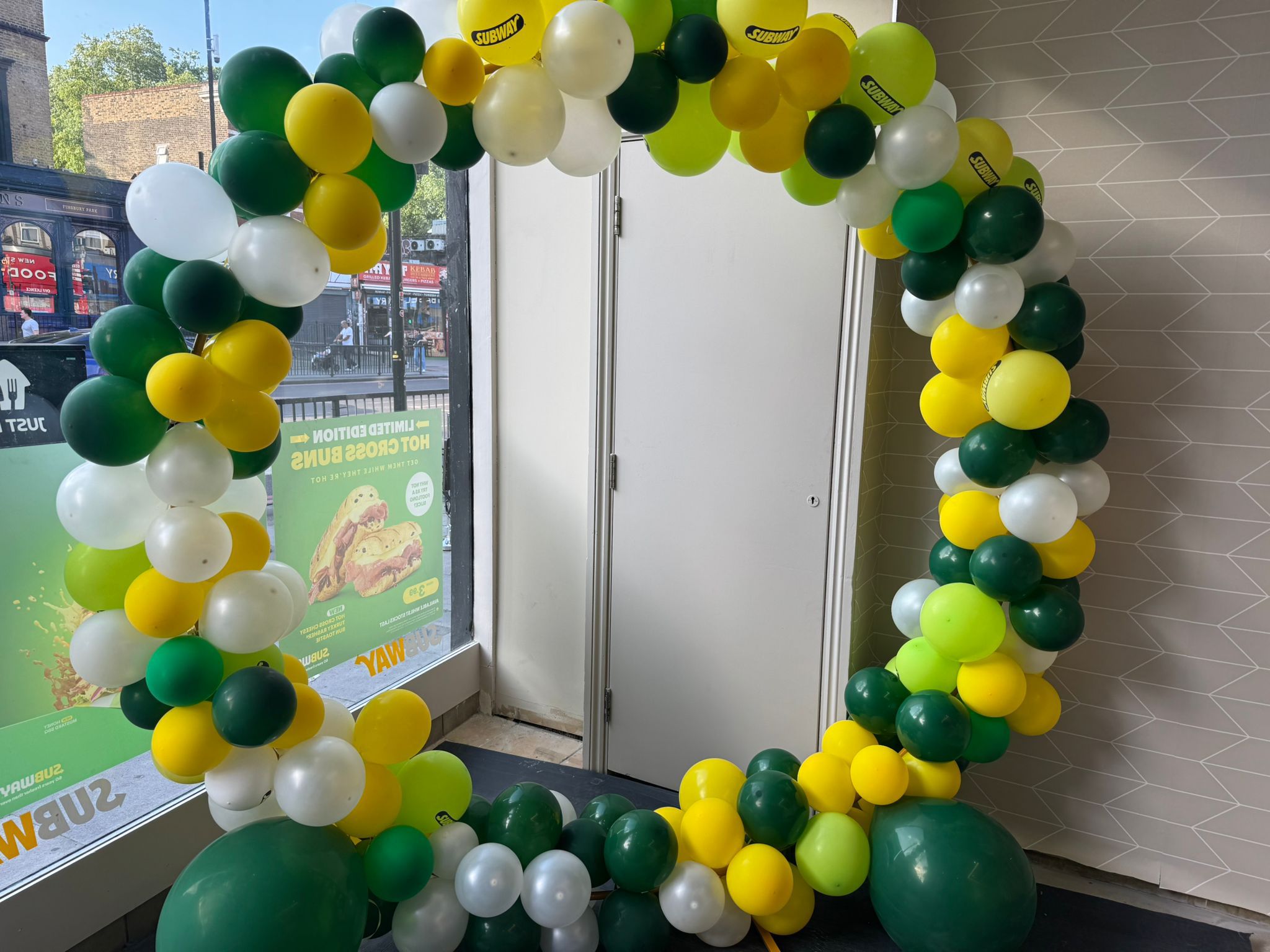 Green, yellow, and white balloon arch decorating a Subway shopfront entrance, marking a store opening or celebration.
