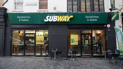 Exterior of a Subway shopfront with green awning reading Sandwiches and Salads, outdoor seating out front.