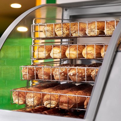 Freshly baked bread rolls displayed on wire racks in a warming case, Subway green wall behind.