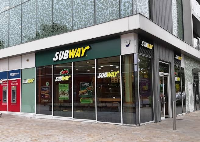 Exterior of a modern glass-fronted Subway store on a street corner, Tesco Bank signage to the left.